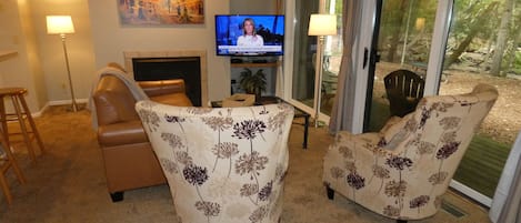 A living room with a fireplace, two floral armchairs, a television, and a sliding glass door leading to an outdoor area.
