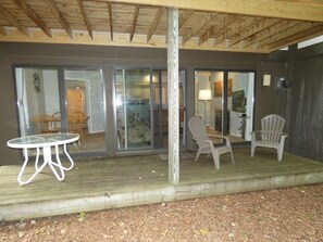 An outdoor covered patio area with a table and two chairs, a wooden deck, and a view into a room with a fireplace. - Whispering Creek at Pinebrook (Bellaire)
