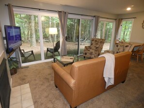 Ein Wohnzimmer mit einer Couch, Sesseln, einem Fernseher und Blick auf einen Wald durch große Schiebeglastüren.