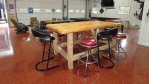Uma oficina com uma longa mesa de madeira, quatro banquinhos e diversas ferramentas e equipamentos.