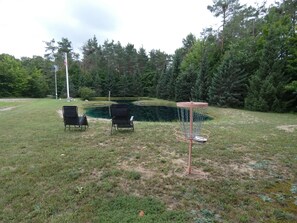 Uma área gramada com um cesto de disc golf, duas cadeiras e um mastro.