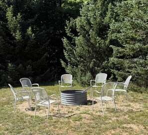 A group of outdoor metal chairs arranged around a circular metal table in a grassy area with dense coniferous trees in the background.