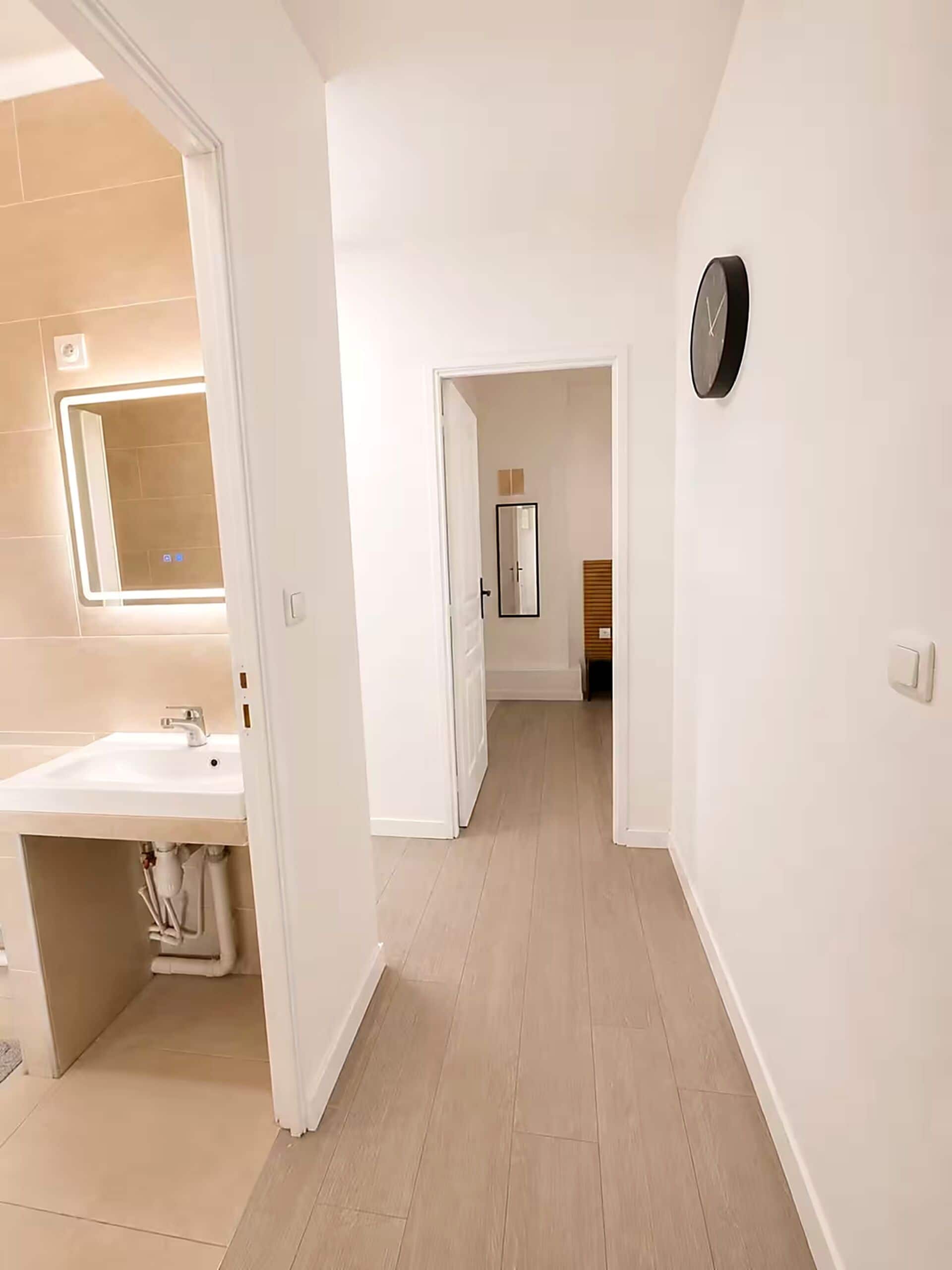 Un corridoio con un bagno a sinistra, uno specchio e una sveglia a destra.