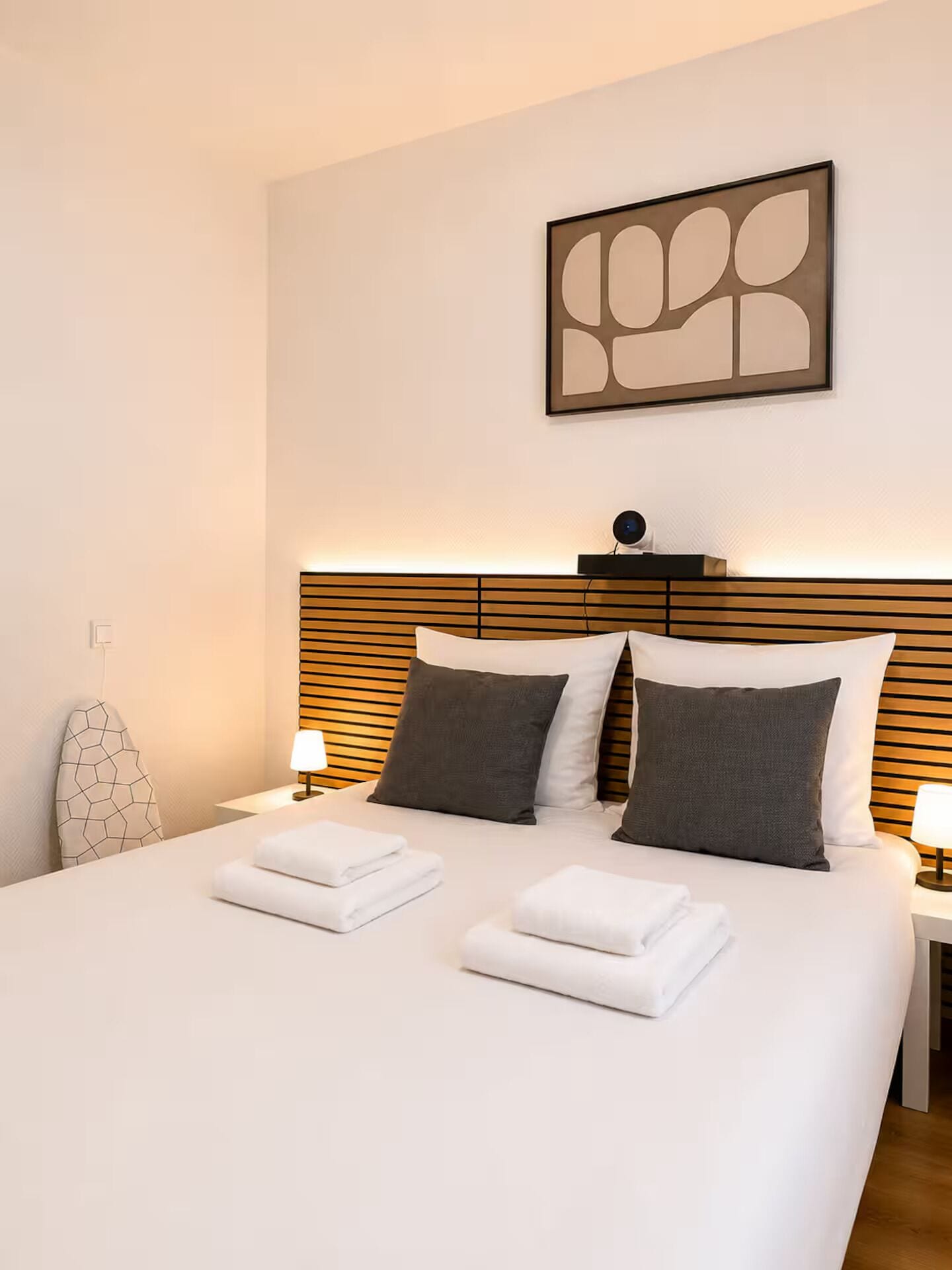 Une chambre d’hôtel avec un lit parfaitement fait, deux lampes de chevet et un tableau abstrait moderne accroché au mur.