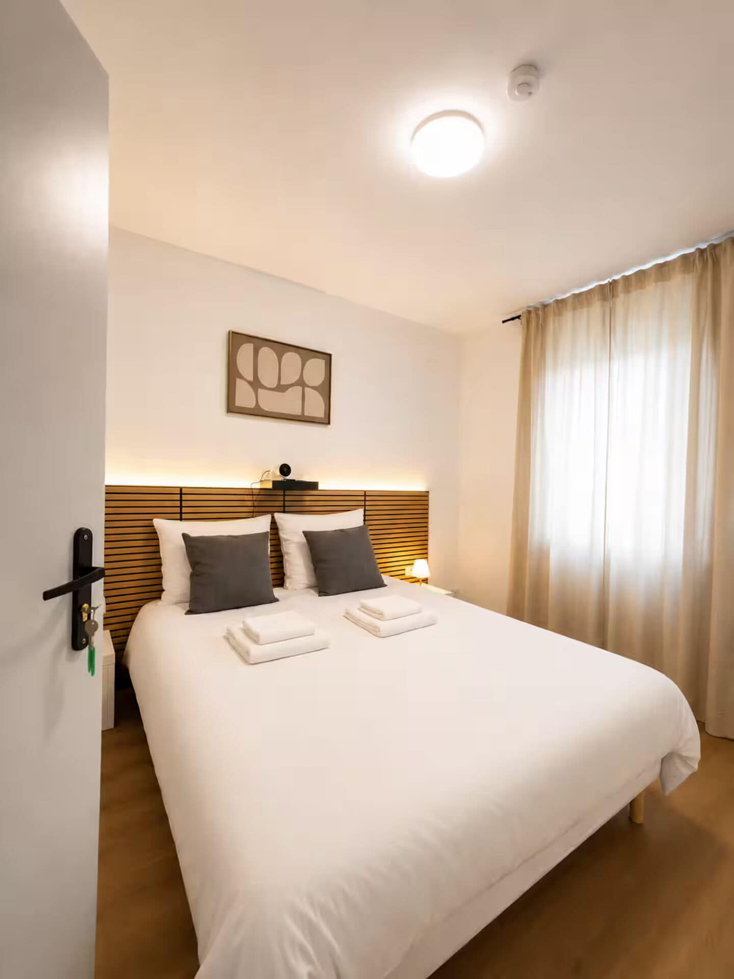 Habitación de hotel con cama, cabecero, mesita de noche, lámpara, una obra de arte en la pared y una ventana con cortinas.