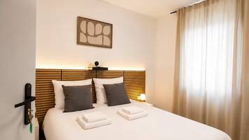 Habitación de hotel con cama, cabecero, mesita de noche, lámpara, una obra de arte en la pared y una ventana con cortinas.