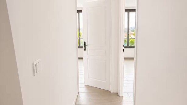 A narrow hallway with white walls and wooden flooring leading to a door.