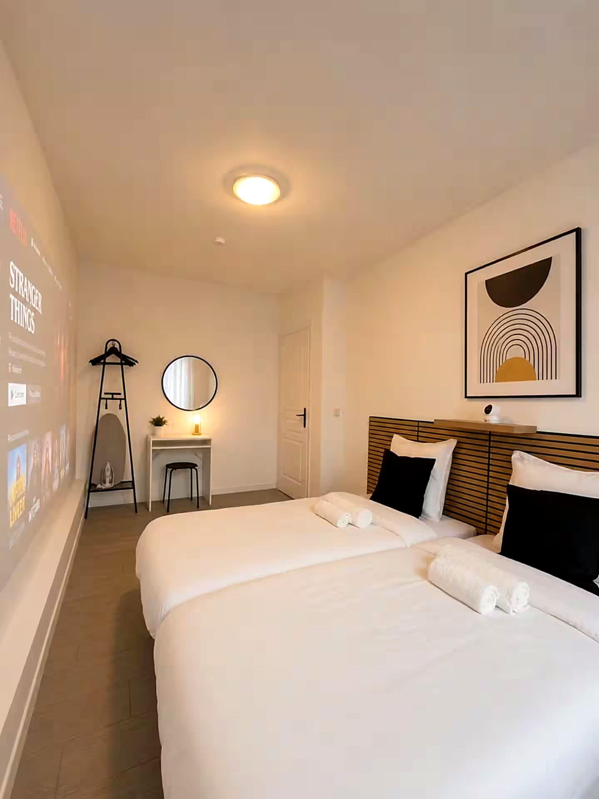 Een hotelkamer met twee bedden, een bureau met stoel, een spiegel en een lijst met een foto aan de muur.