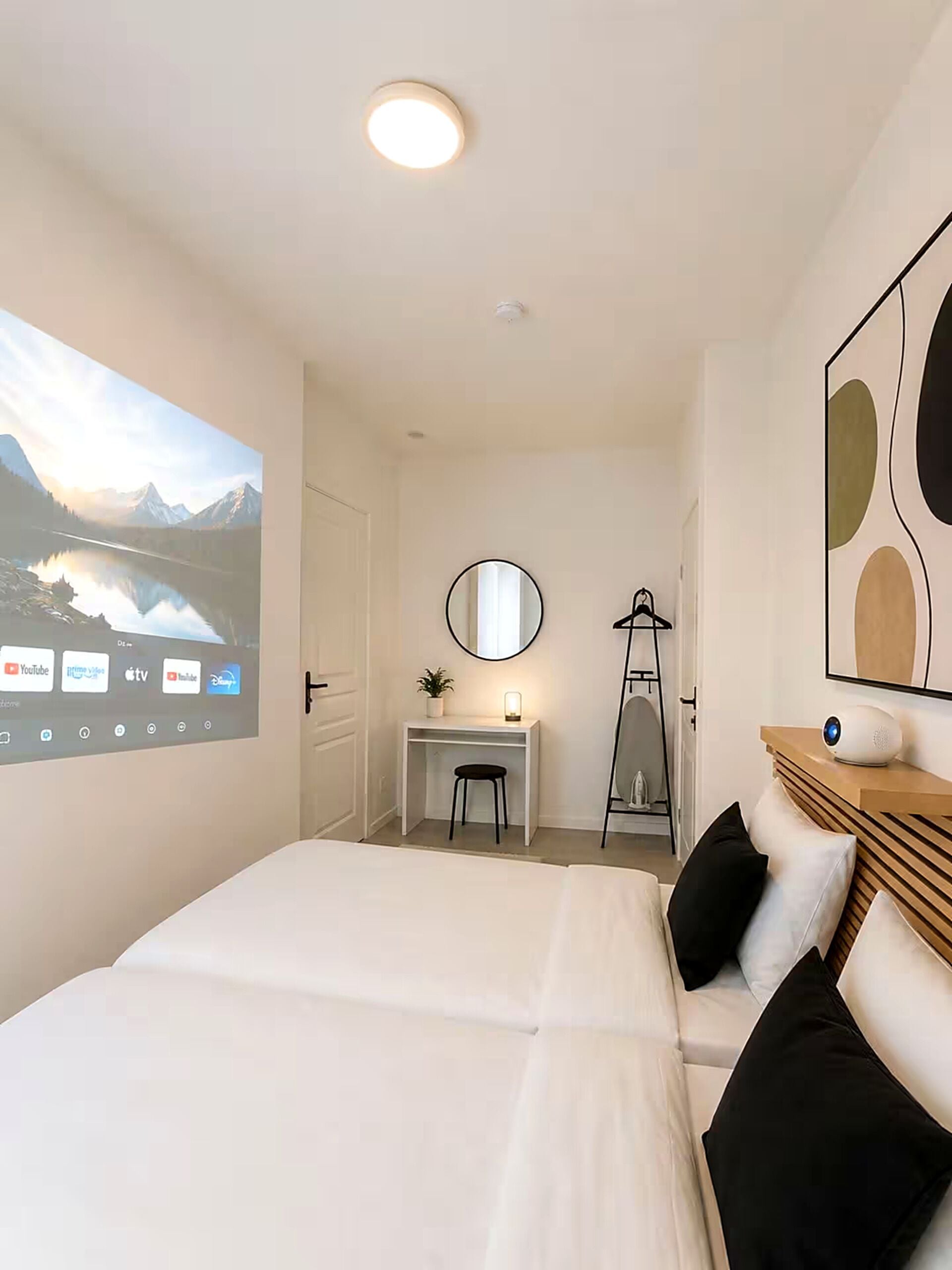 Une chambre à coucher comprenant un lit, un bureau, une chaise, un miroir et un écran de projection sur lequel est diffusé un paysage pittoresque.