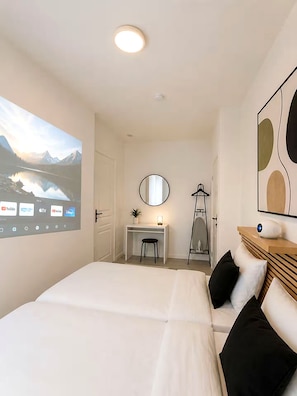 A bedroom with a bed, a desk, a chair, a mirror, and a projector screen displaying a scenic landscape. - Hôtel de la gare (Pantin)