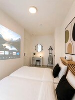 Un dormitorio con cama, un escritorio, una silla, un espejo y una pantalla proyectando un paisaje pintoresco.