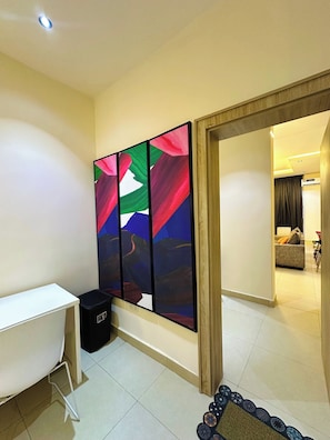 Um quarto com uma escrivaninha branca, um cesto de lixo preto e um quadro abstrato colorido na parede.