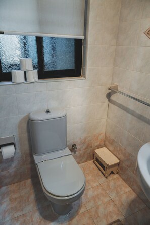 A bathroom with a toilet, a small wooden stool, a window, and a towel dispenser. - Charming 3-Bedroom Apartment with 3 Terraces (San Pawl il-Baħar)