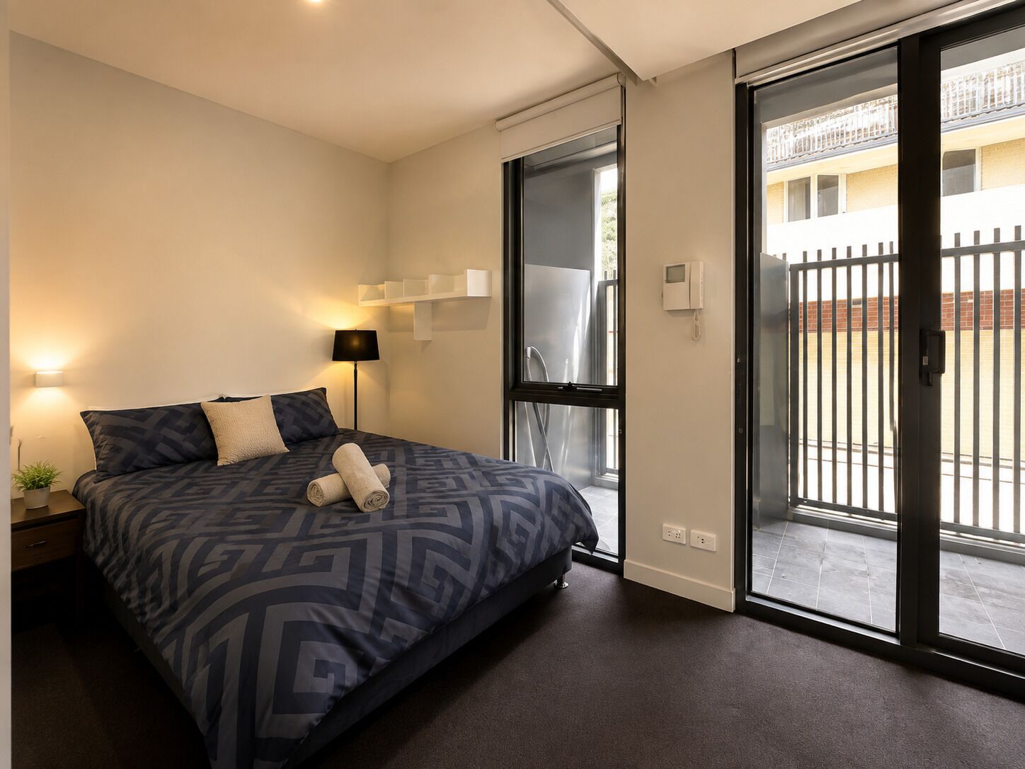 A modern bedroom with a large bed, a bedside lamp, a small table with a plant, and a sliding glass door leading to an outdoor area.
