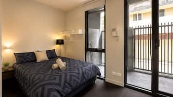A modern bedroom with a large bed, a bedside lamp, a small table with a plant, and a sliding glass door leading to an outdoor area.