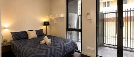 Un dormitorio moderno con una cama grande, una lámpara de noche, una mesita con una planta y un ventanal corredero que da a un área exterior.