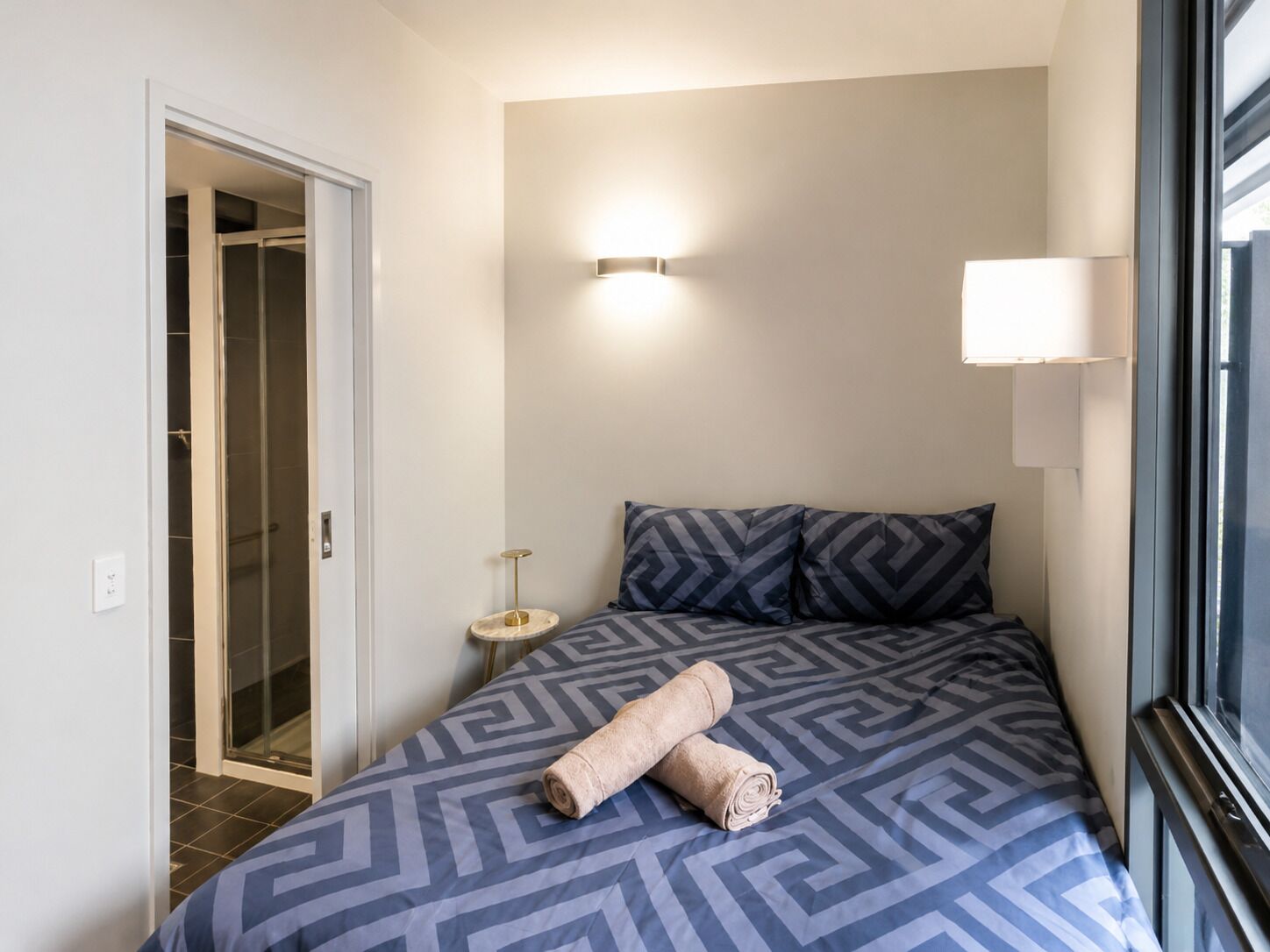 A hotel room with a bed, a bedside lamp, a small table, and a window.