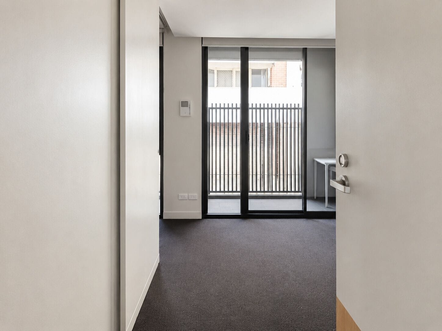A hallway leading to a balcony with a metal railing, a small table, and a door with a handle.