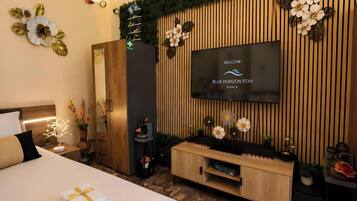 Una camera d'albergo con un letto, una TV a parete, un mobiletto in legno e un tavolino con un vaso di fiori.