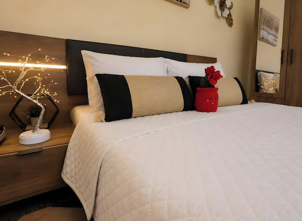 Una cama bien hecha con colcha blanca, cabecera de madera y mesita de noche con una lámpara decorativa con forma de árbol.