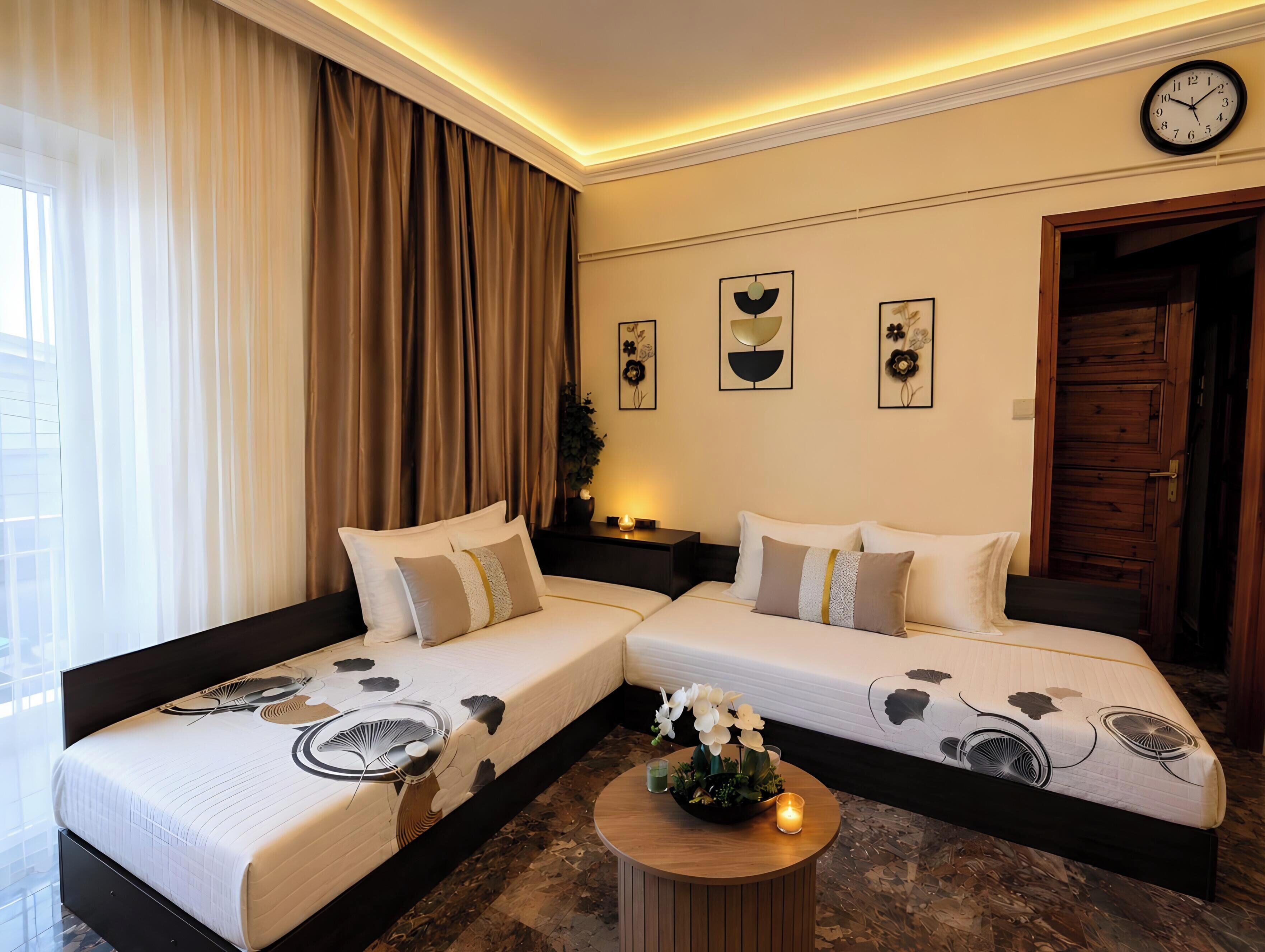 Une chambre d’hôtel avec deux lits, une table ronde en bois sur laquelle est posée une bougie, et une horloge au mur.