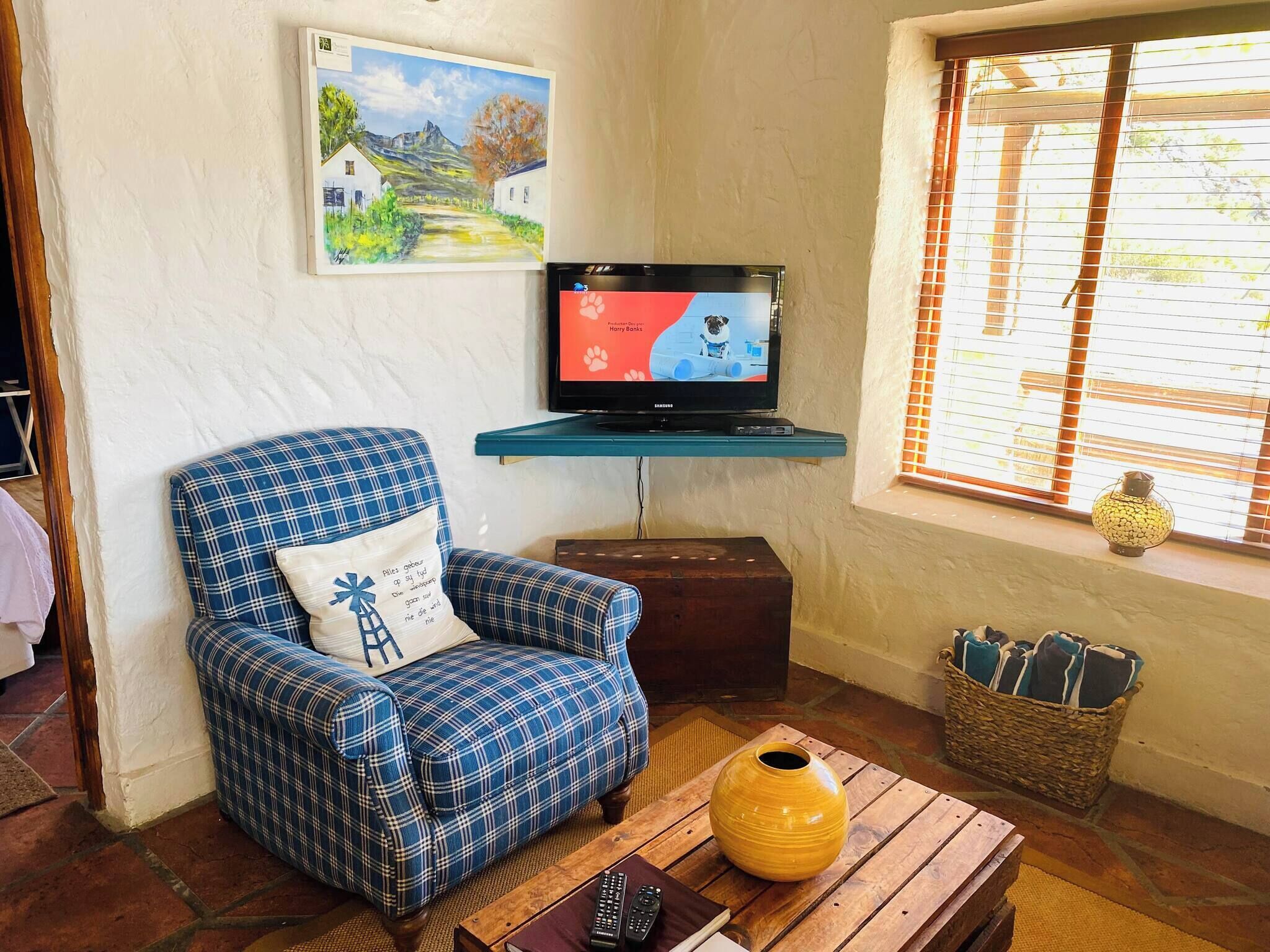Una sala de estar con un sillón a cuadros, un televisor sobre un mueble bajo, una ventana con persianas, un florero sobre una mesa y un cuadro en la pared.