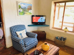 Una sala de estar con un sillón a cuadros, un televisor sobre un mueble bajo, una ventana con persianas, un florero sobre una mesa y un cuadro en la pared.
