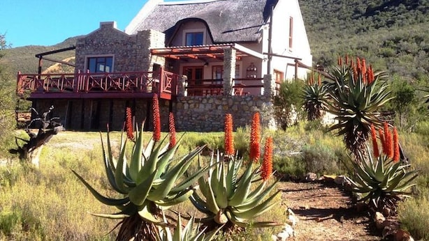 Uma casa de dois andares com fachada de pedra, varanda com corrimão vermelho e montanhas ao fundo.