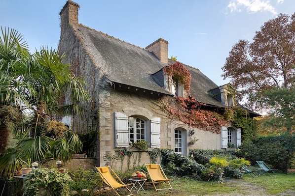 A stone house with a sloped roof, ivy-covered walls, and a garden with two sun loungers.