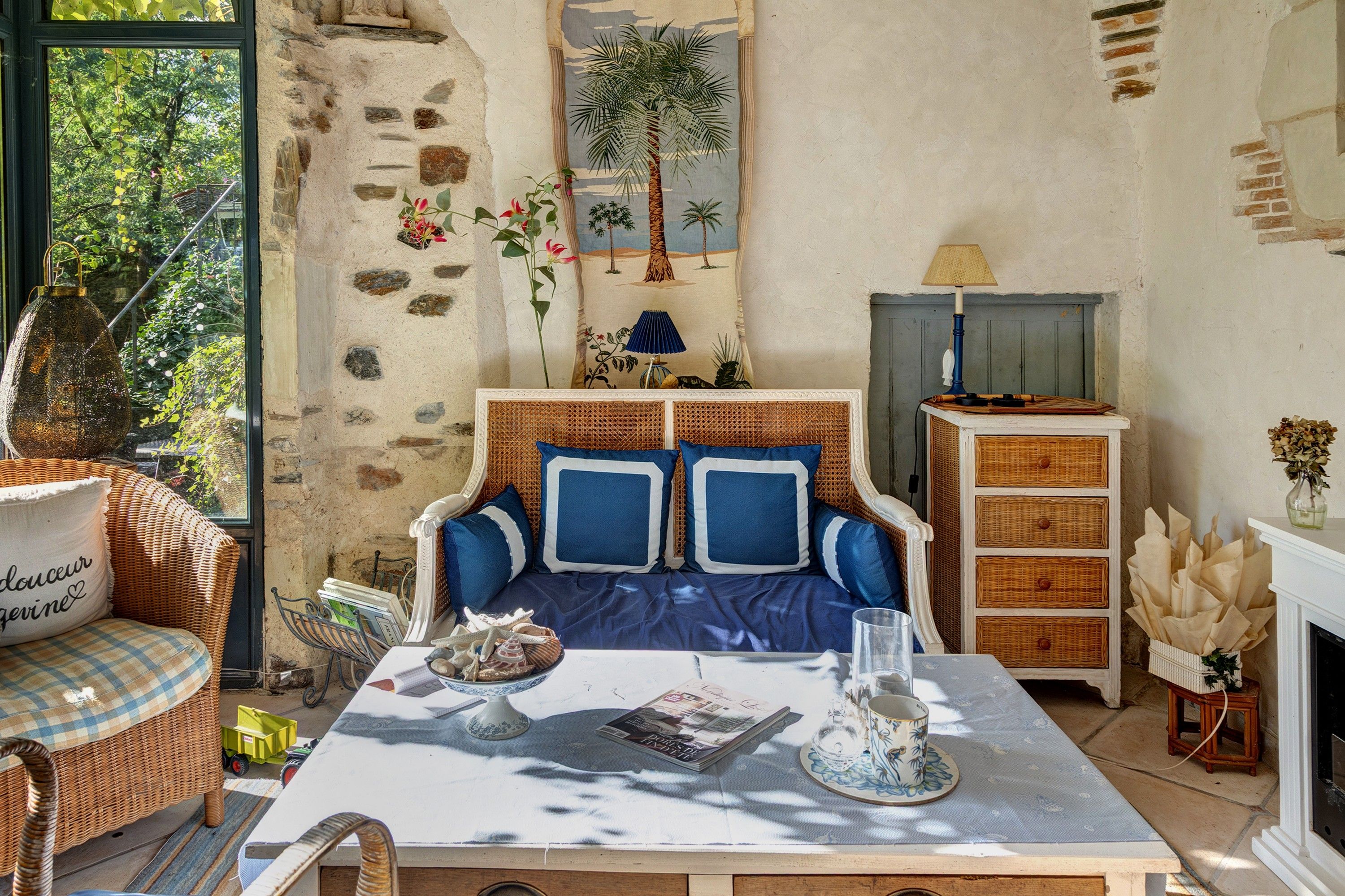 Een knusse woonkamer met een stenen muur, een blauwe bank, rieten meubels en een open haard.