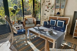 A cozy room with a wicker chair, a sofa with blue cushions, a table with a book and a glass, and a view of a garden through a window.