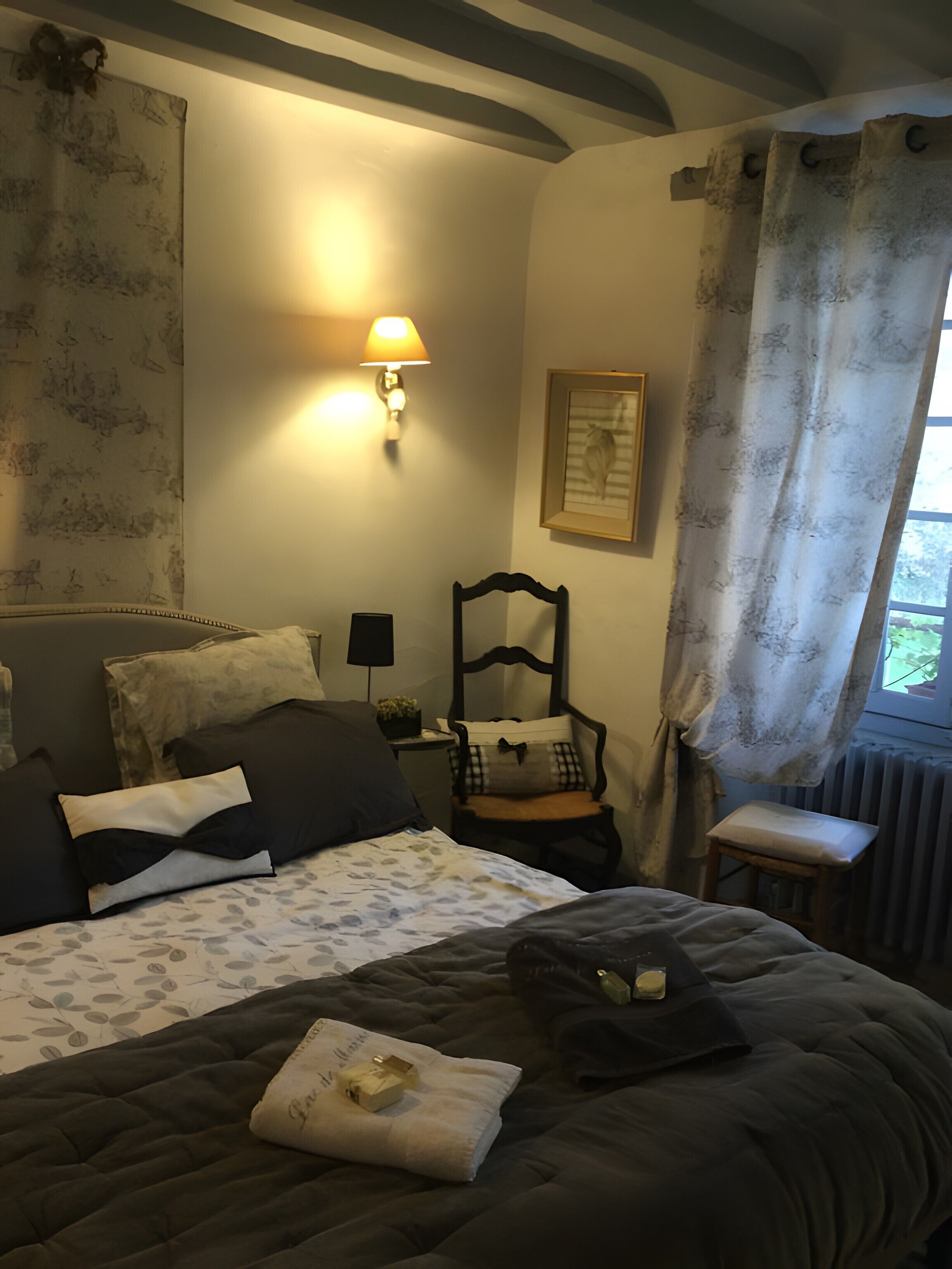 A bedroom with a bed, a chair, a nightstand, a lamp, and a window with curtains.