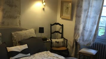 Un dormitorio con cama, una silla, una mesita de noche, una lámpara y una ventana con cortinas.