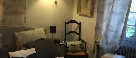Un dormitorio con cama, una silla, una mesita de noche, una lámpara y una ventana con cortinas.