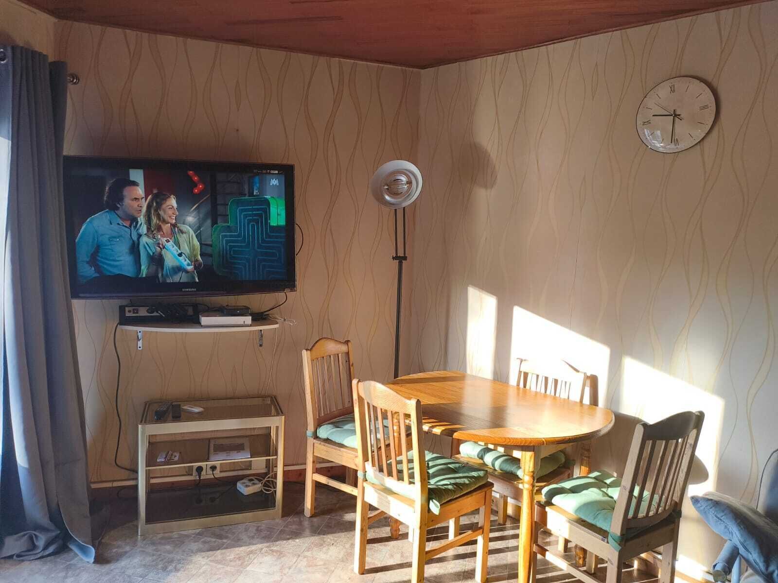 A living room with a television, wooden dining table, chairs, and a clock on the wall.