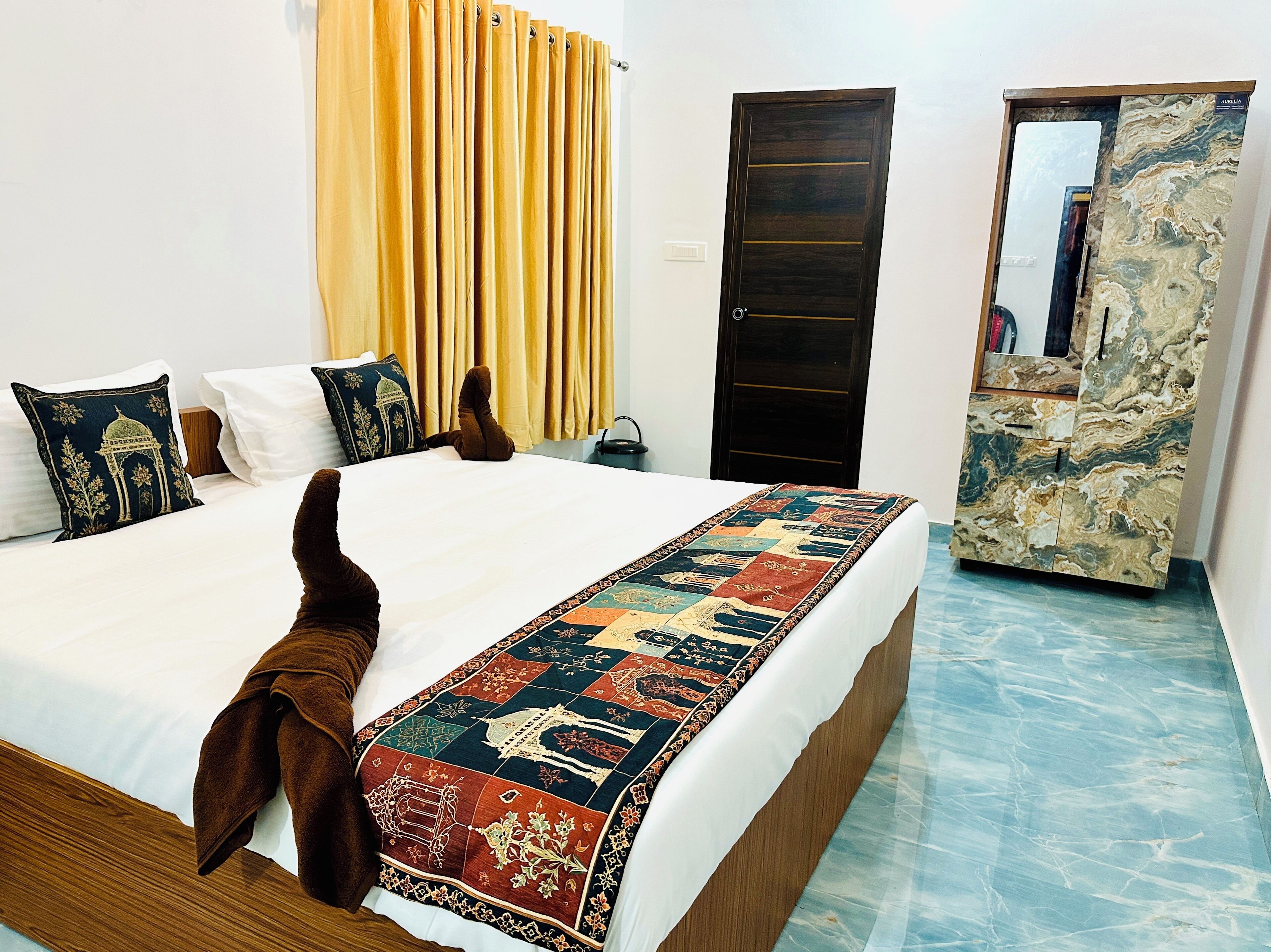 Hotelkamer met een bed, een houten hoofdbord, een beddekleed met dessin, een paar bruine sloffen, een geel gordijn, een donker houten deur en een kast met een marmerlook.