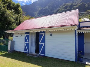 A small building with a red roof and blue doors, surrounded by greenery and mountains.