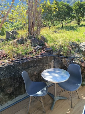 Un espacio al aire libre con dos sillas y una mesa, rodeado por un muro de piedra y vegetación.