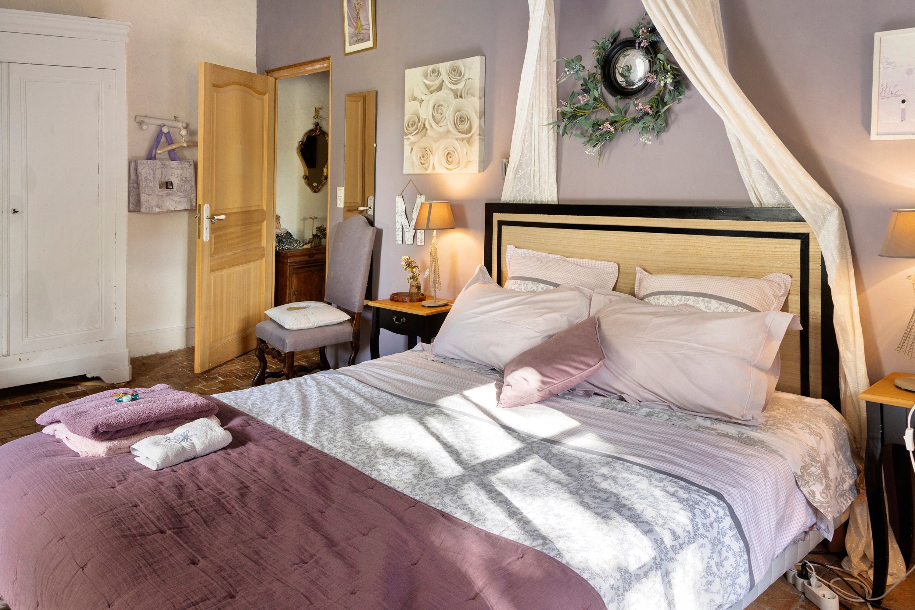 A bedroom with a bed, a bedside table, a nightstand, a mirror, a door, and a wall with a floral decoration.