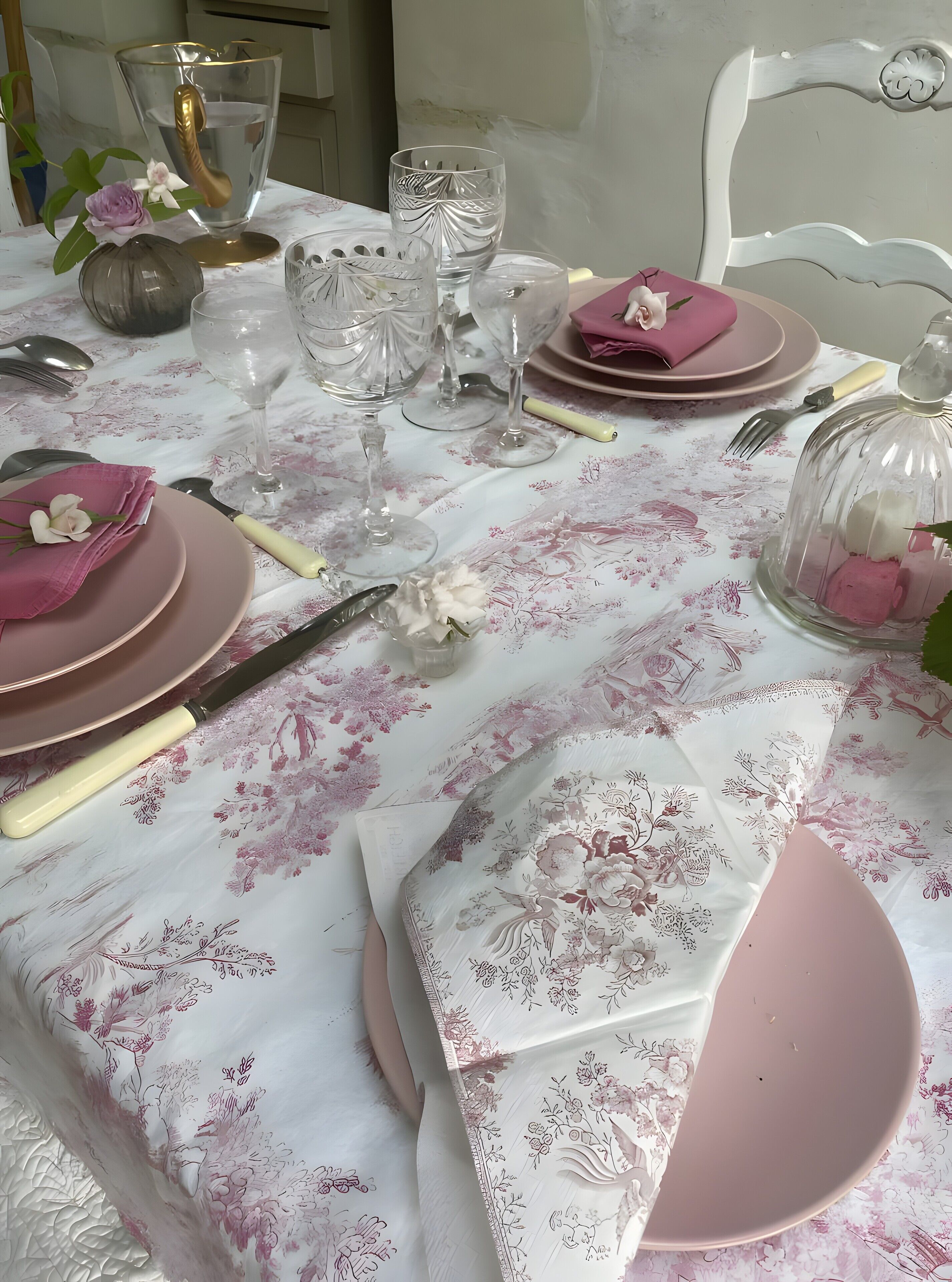 Una mesa dispuesta con platos rosas, copas transparentes y un mantel con motivos florales.