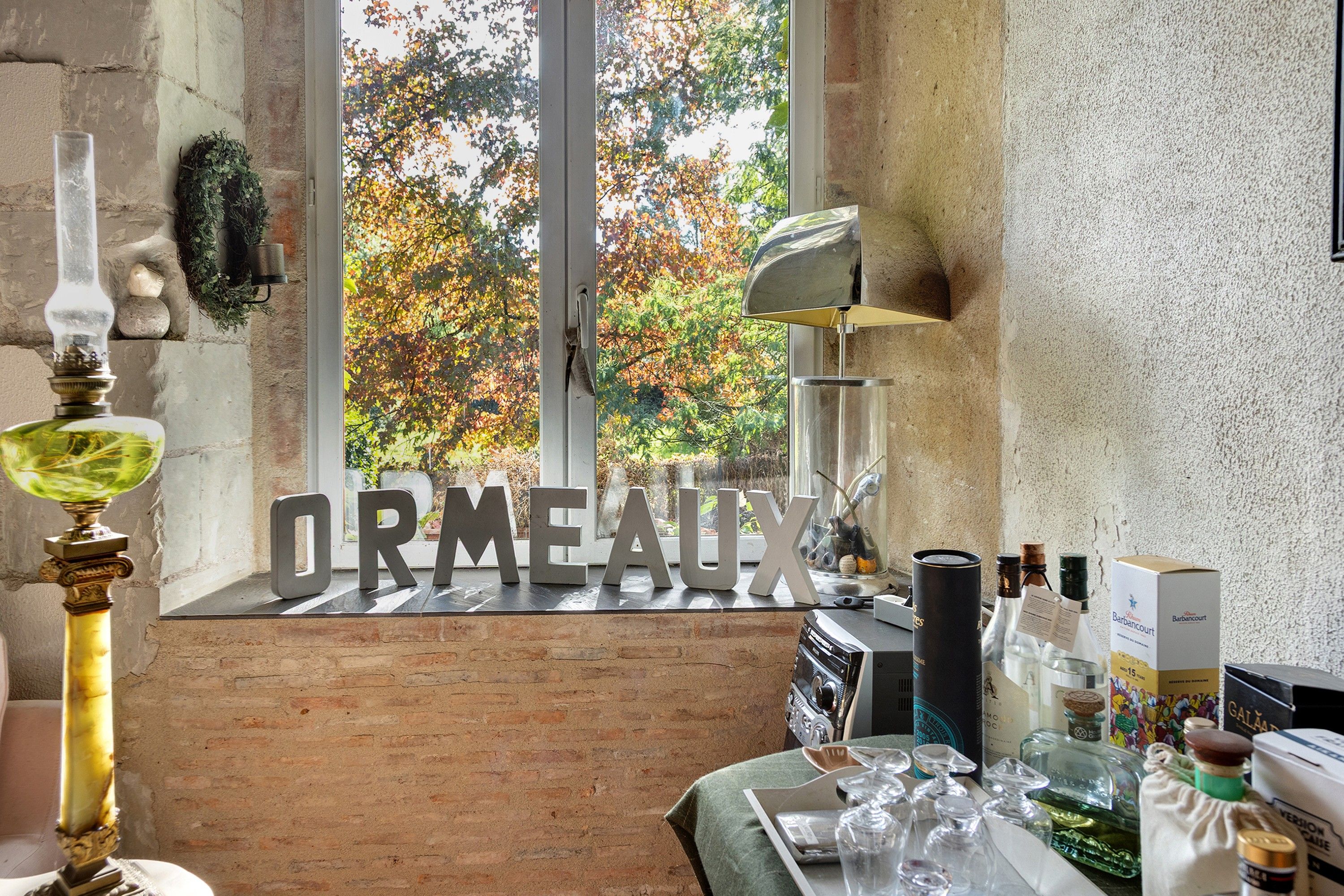 Een gezellige kamer met een groot raam, een decoratief bord waarop 'ORMEAUX' staat, en diverse flessen en glazen op de vensterbank.