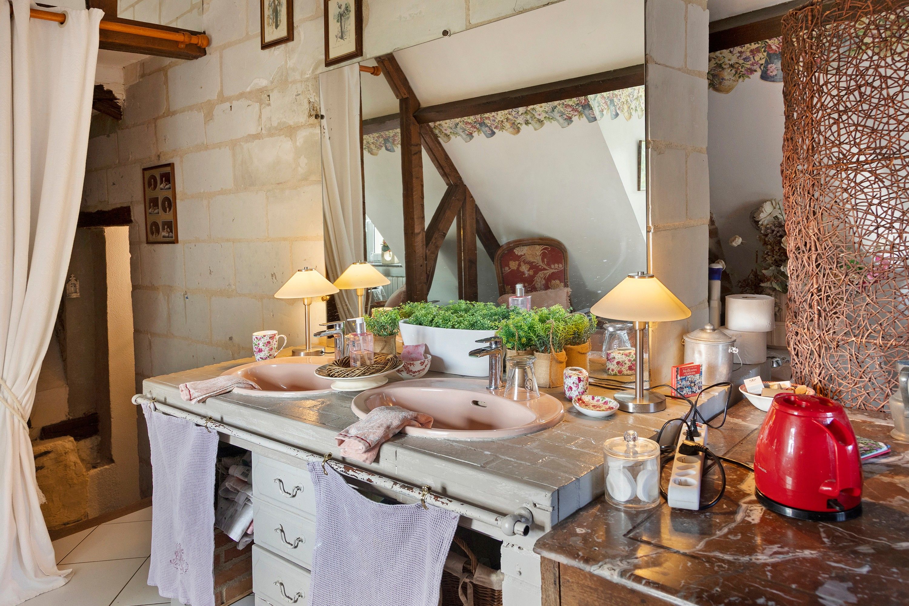 A bathroom with a double sink, two wall-mounted lamps, a red kettle, and a mirror above the sink.