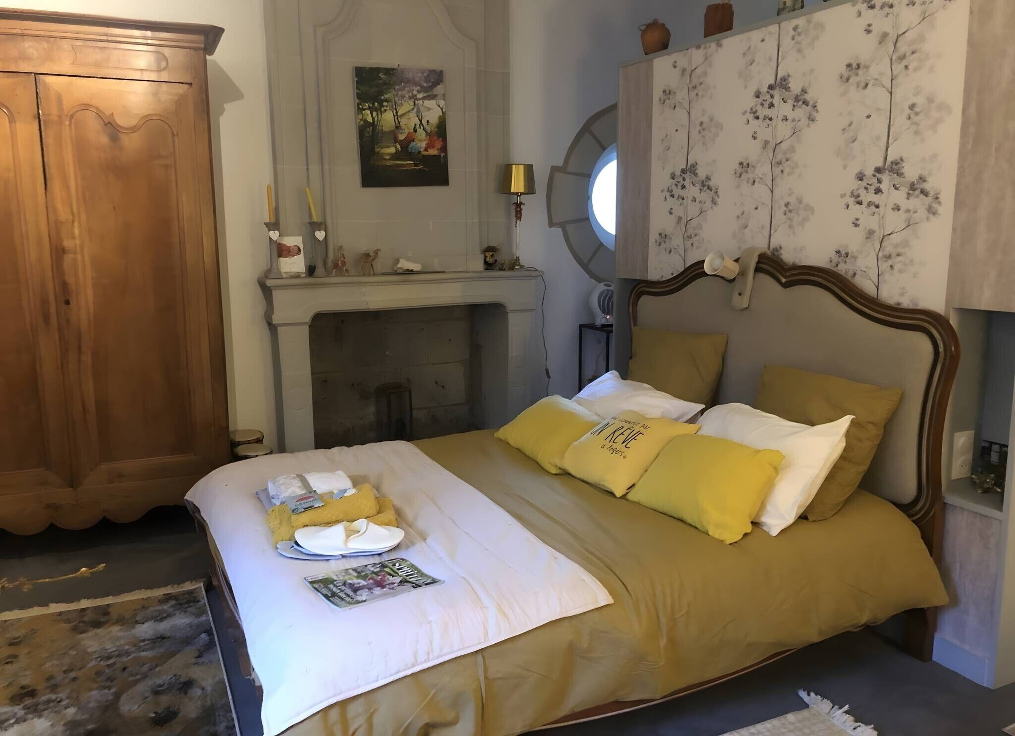 A neatly made bed with a yellow and white color scheme, a wooden wardrobe, a fireplace, and a painting on the wall.