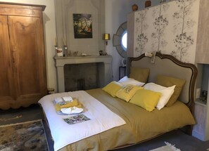 A neatly made bed with a yellow and white color scheme, a wooden wardrobe, a fireplace, and a painting on the wall.