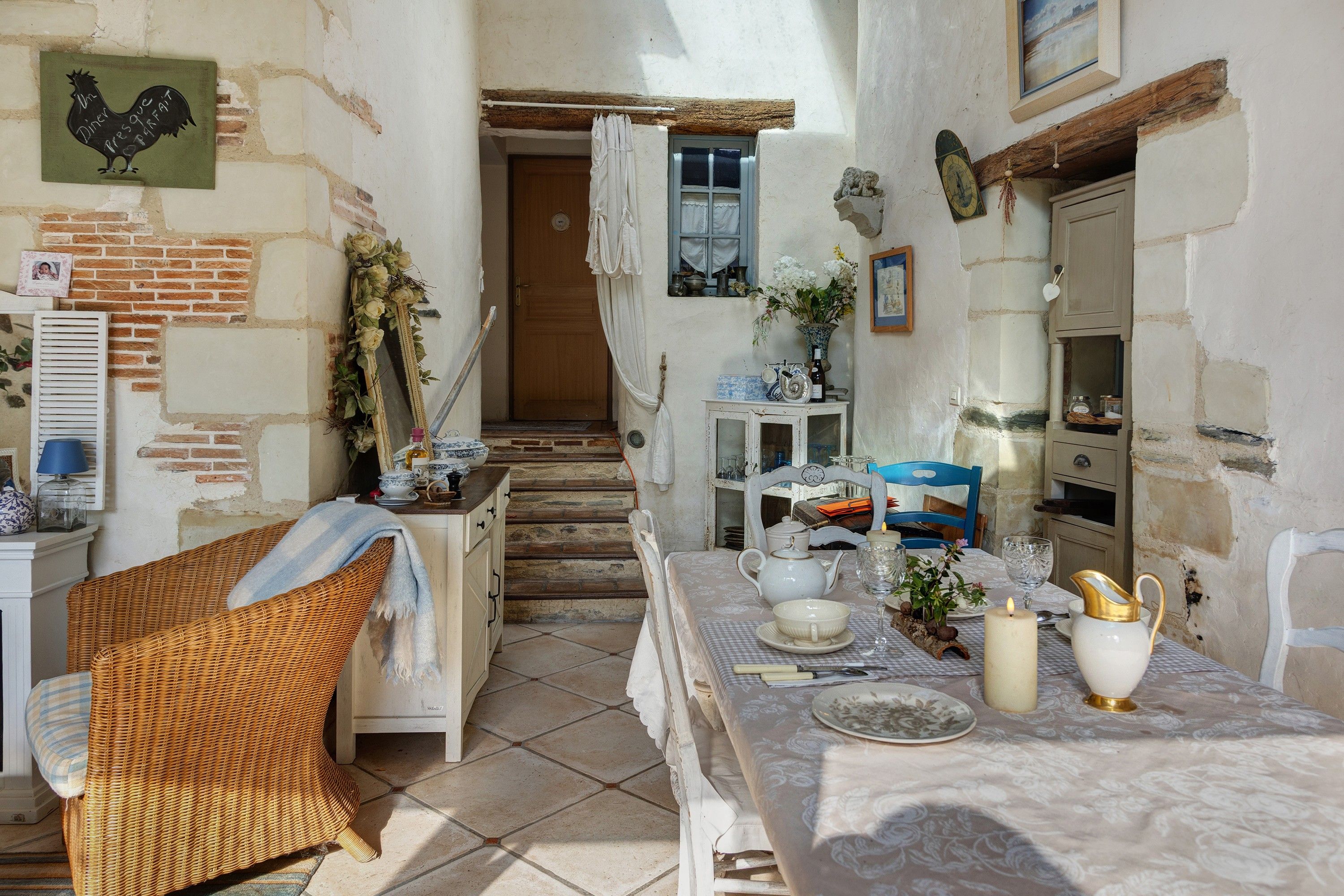 A rustic dining area with a table set for two, a wicker chair, and a staircase leading to a door.