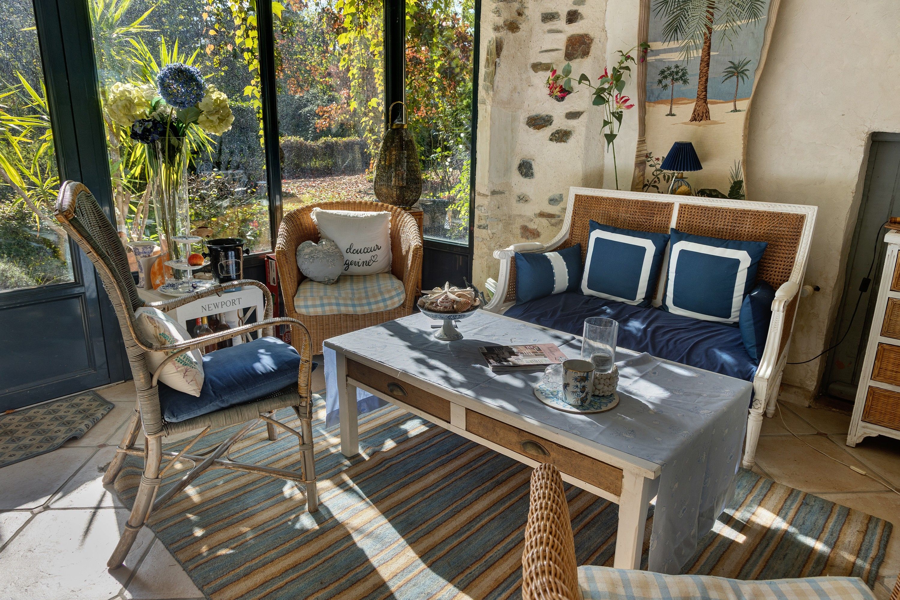 A cozy room with a wicker chair, a sofa with blue cushions, a table with a book and a glass, and a view of a garden through a window.