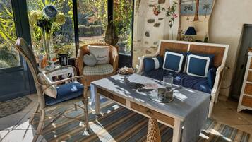 A cozy room with a wicker chair, a sofa with blue cushions, a table with a book and a glass, and a view of a garden through a window.