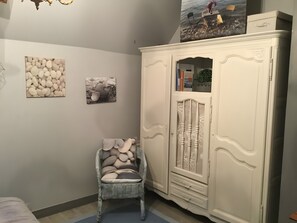 A room with a white wardrobe, a wicker chair with cushions, and two wall-mounted pictures. - Éole – Family House with Shared Terrace, Private Garden, and Wi-Fi (Cancale)