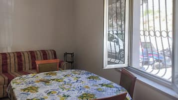 Une pièce avec une table ronde recouverte d’une nappe à motifs floraux, un canapé à rayures et une fenêtre ornée d’un grillage métallique.