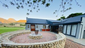 A circular stone patio with a fire pit, a building with a blue roof, and a view of mountains in the background. - Spacious Farmhouse Rental in Ladismith With Stunning Views (Ladismith)